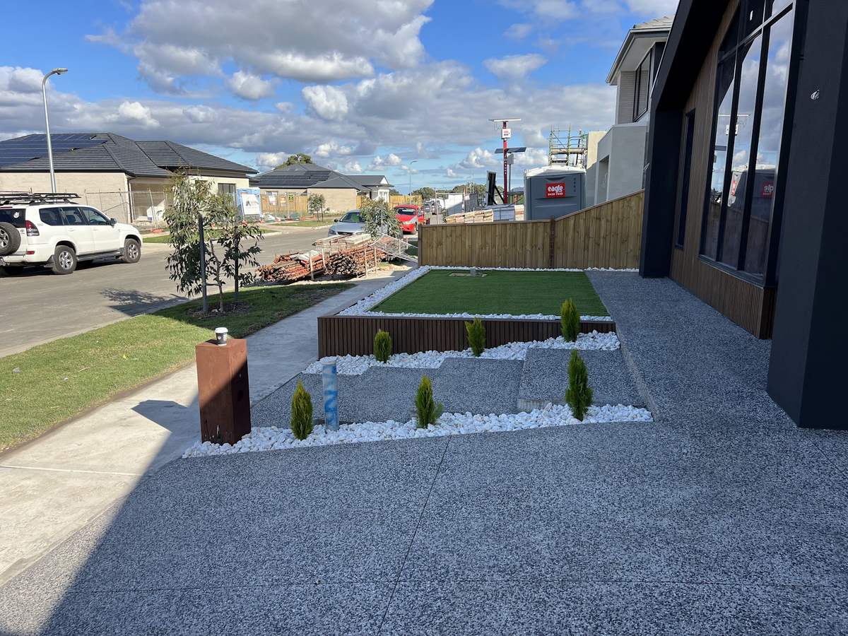 A finished front yard with raised turf bed, white pebbles, pencil pines and exposed aggregate in front of a dark home