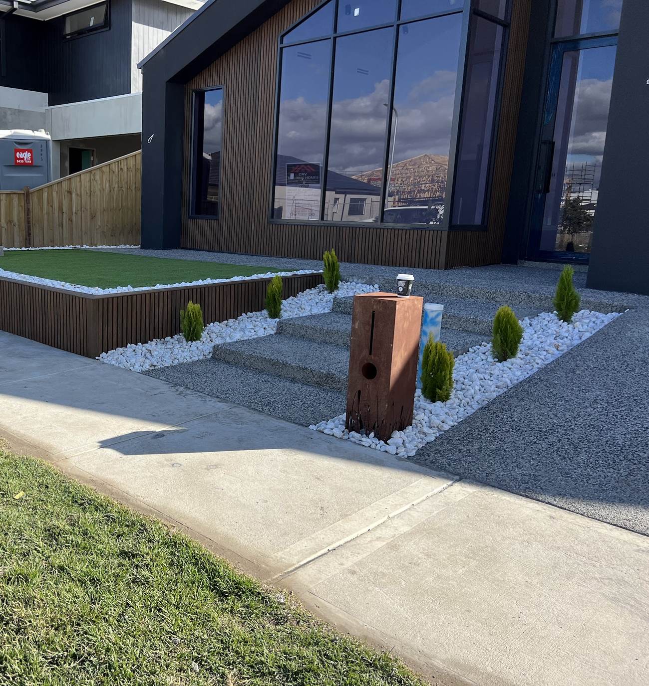 A finished front yard — raised turf terrace with slatted timber cladding, exposed aggregate steps and a corten letterbox in front of a dark architectural home