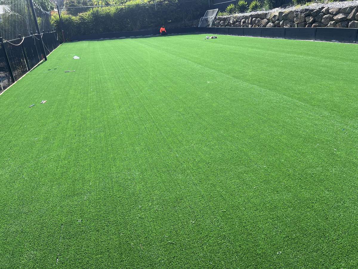 A full-size synthetic tennis court with white lines, set beneath a boulder retaining wall